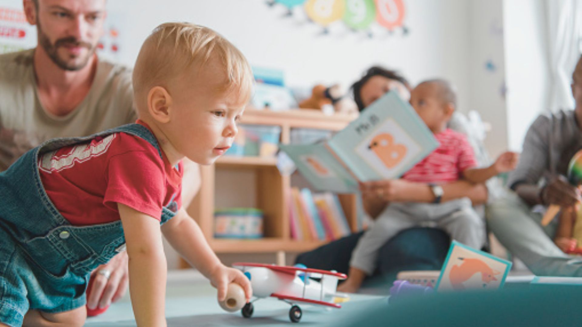 A baby in red shirt and denim overalls crawls toward a toy airplane in a playroom.