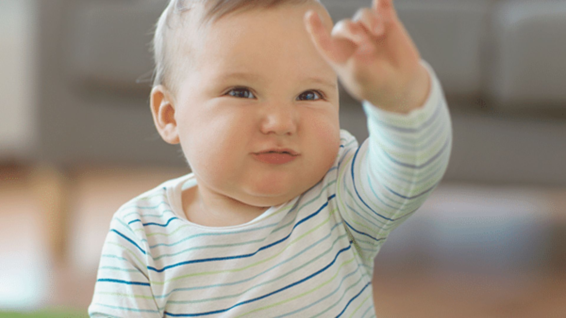 A smiling baby in a striped onesie sits indoors and raises one hand with eyes focused upward.