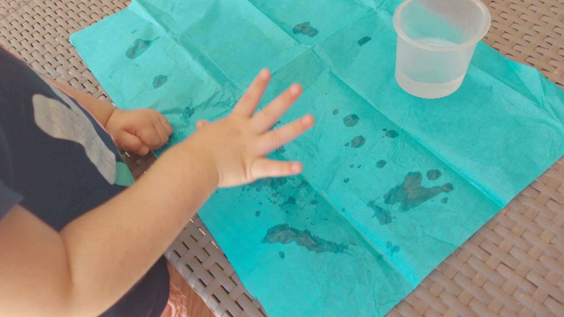 An infantโs hand trying to draw on a blue tissue paper with water droplets beside a small cup.