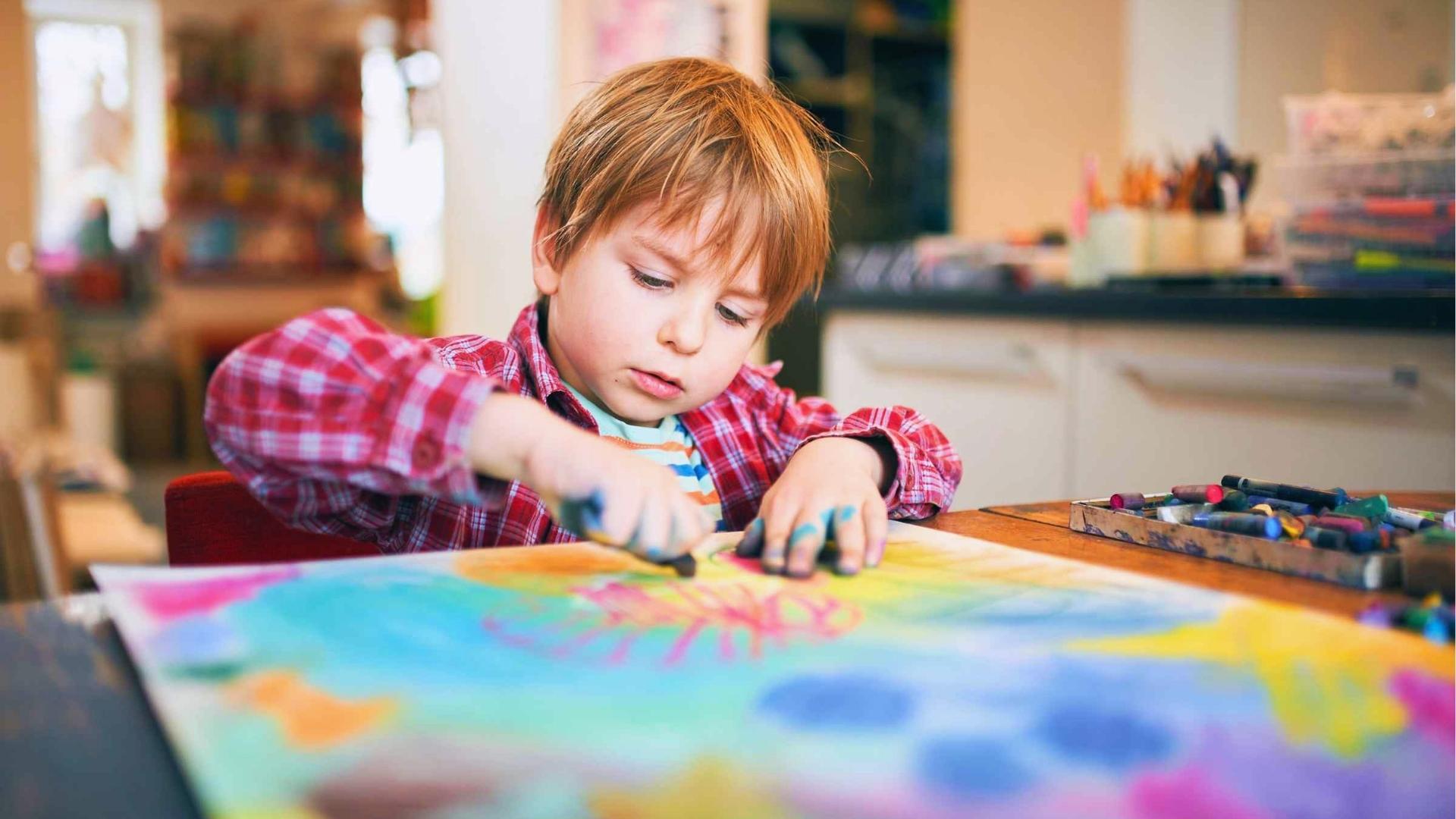 Boy in red plaid draws with pastels at table, hands blue, focused art therapy, Activities for Autistic Kids.