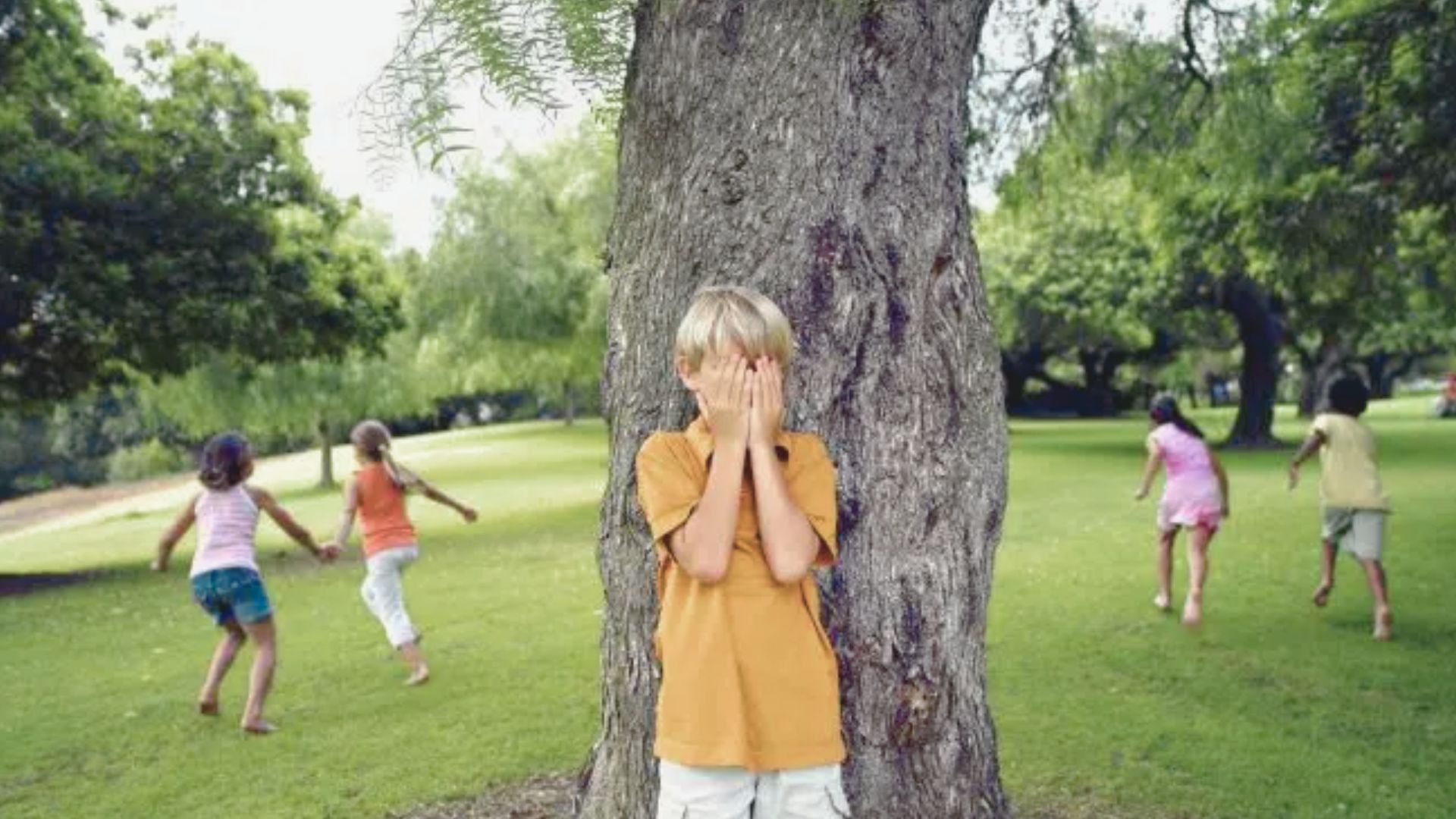 A boy covers his eyes by a tree while other children run across a grassy park.