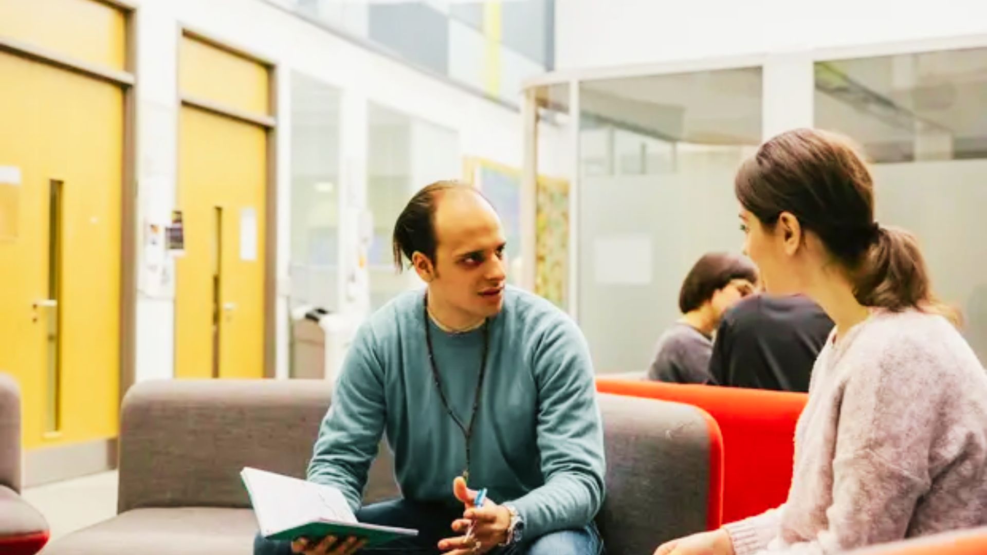 A man and woman sit on couches in a library, surrounded by books, engaged in conversation.
