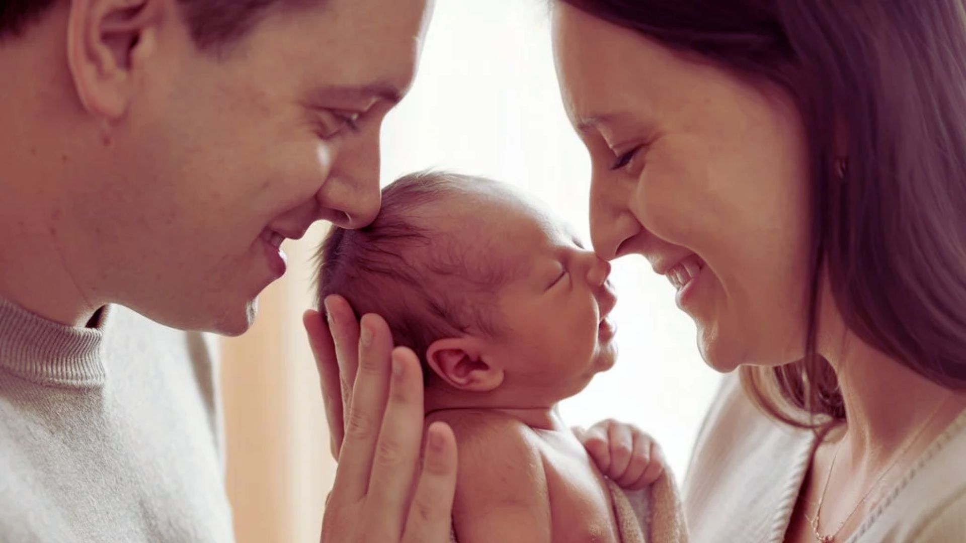 A man and woman gently hold a baby together, smiling at each other in a warm, familial moment.