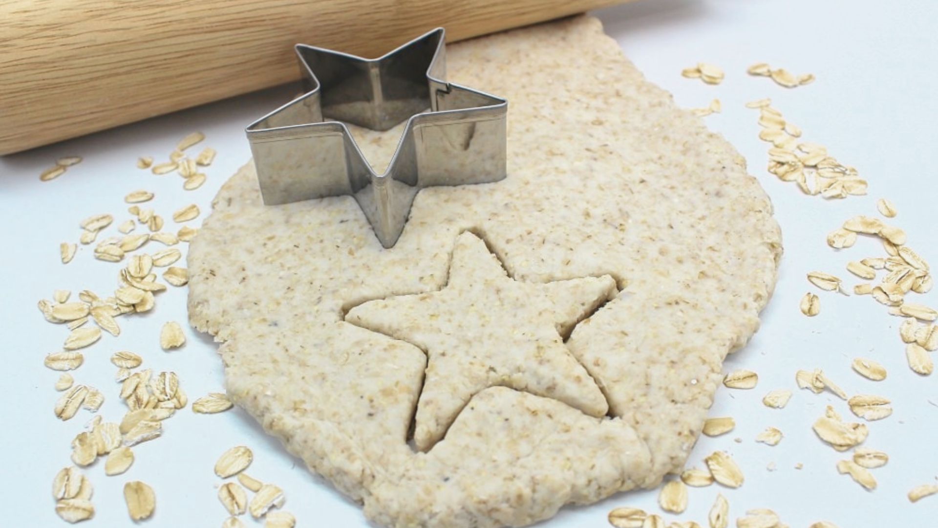 Rolled oat dough sits on a white surface with a star-shaped cookie cutter and scattered oats nearby.