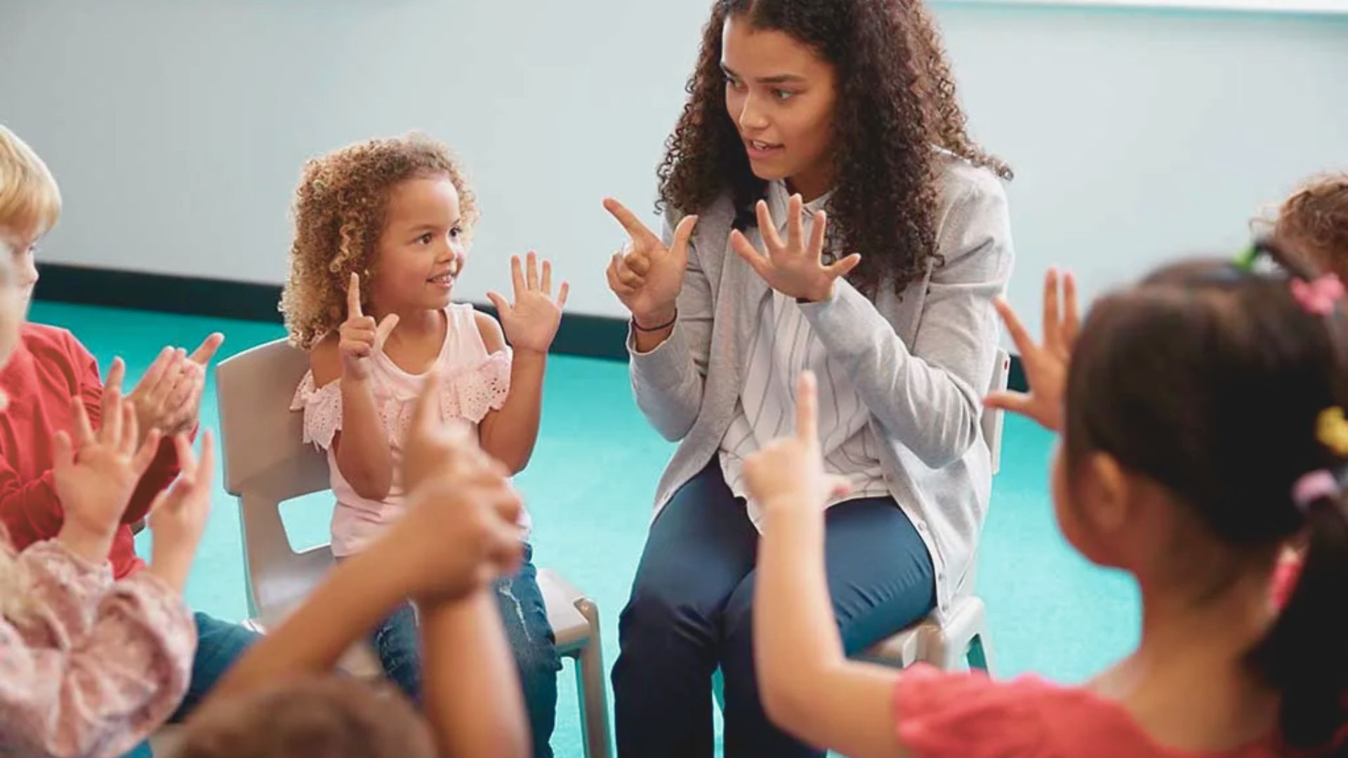 A teacher sits in a circle with children, demonstrating finger and hand movements as they mirror her.
