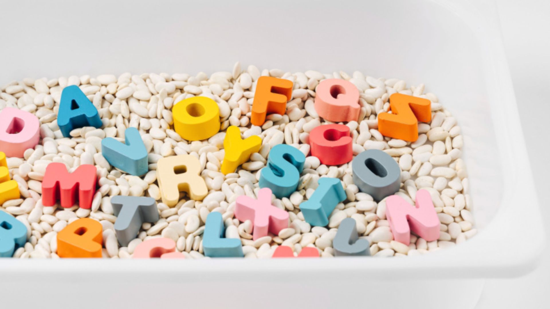 White bin filled with dried white beans and colorful wooden uppercase letters mixed throughout for a tactile alphabet search.