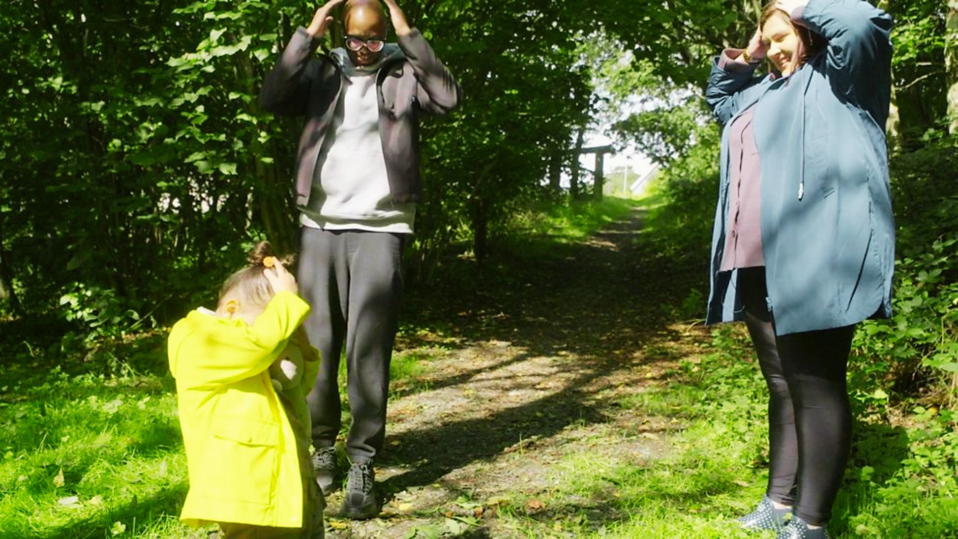 A man and woman stand in the woods with a child, surrounded by tall trees and dappled sunlight filtering through the leaves.