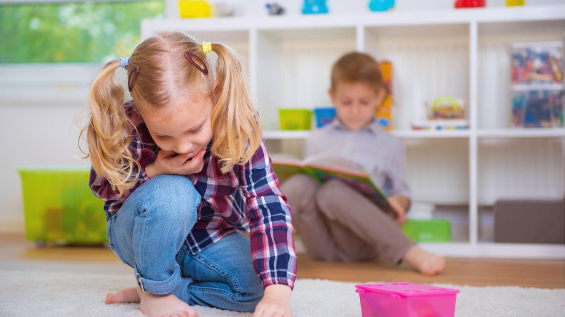 Girl with pigtails plays on floor, boy reads book, toys, bright playroom shelves.