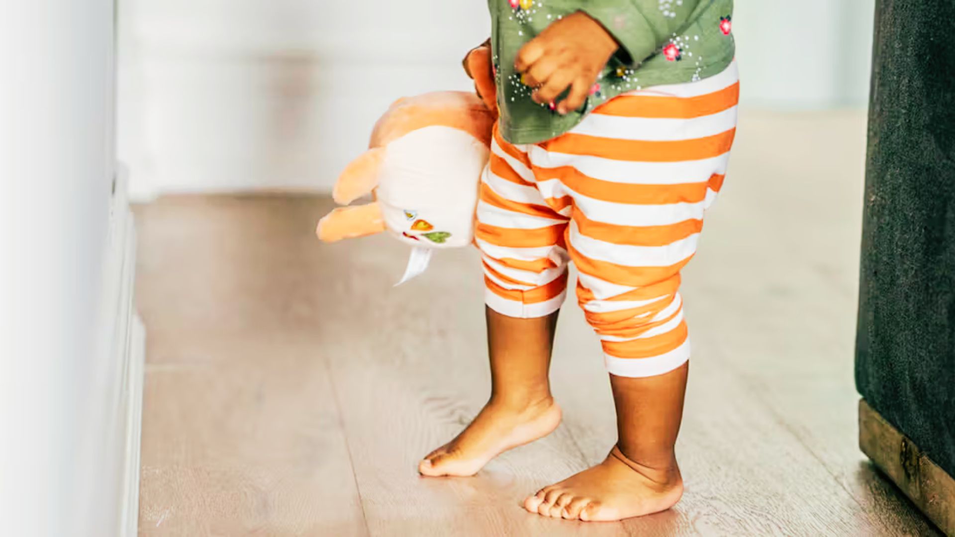 A toddler stands on a wooden floor, holding a stuffed animal in his hand.