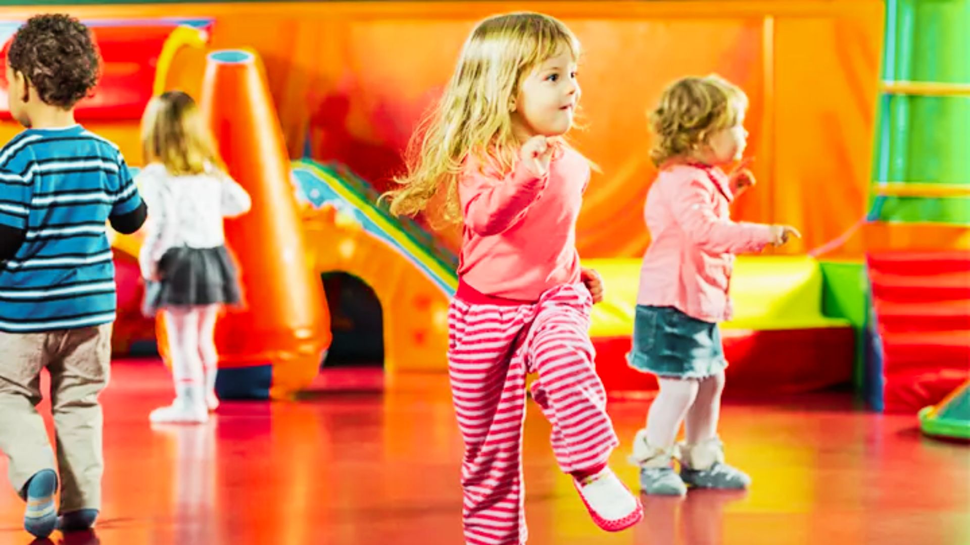 Children joyfully dancing in a colorful indoor play area filled with slides, and soft play equipment.