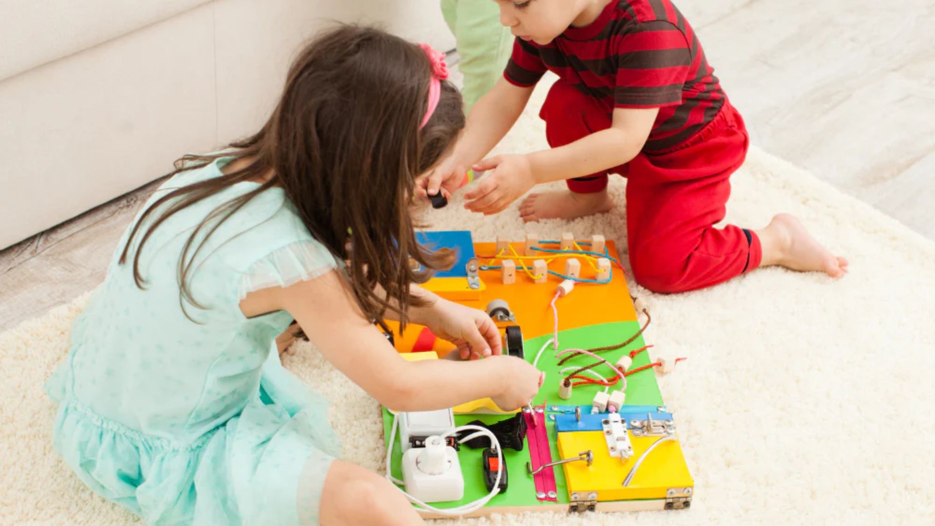 Two toddlers playing together on a carpet with a wooden busy board, developing fine motor skills through a collaborative sensory activity at home
