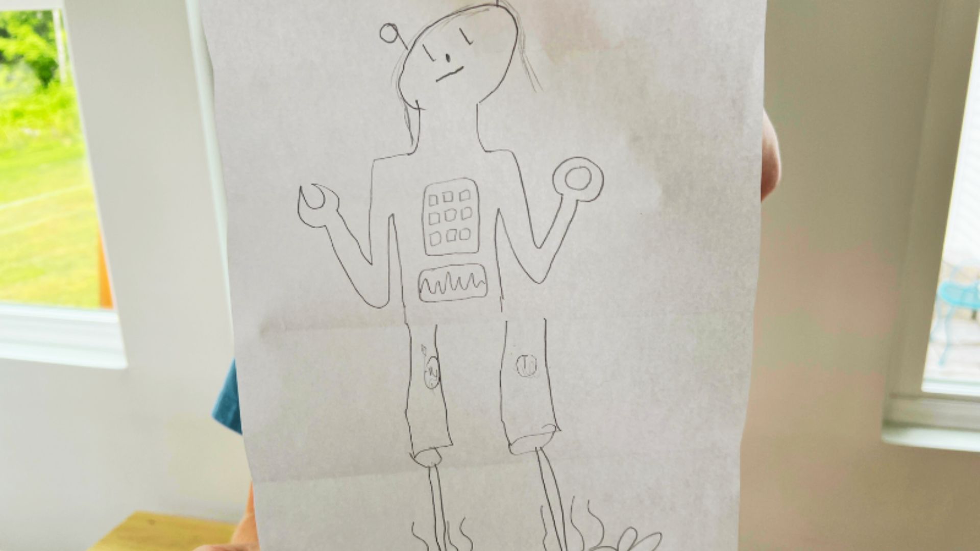 A child proudly displays a drawing of a robot, smiling as they hold it up for others to see.