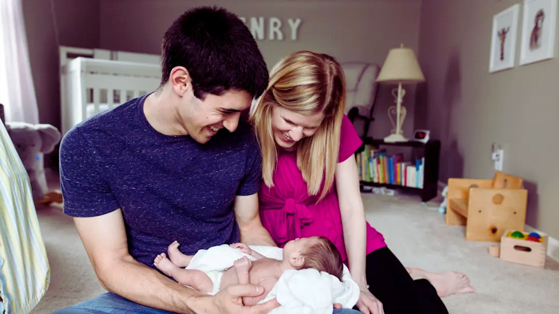 A man and woman hold a baby together in a cozy room, smiling at each other with warmth and affection.