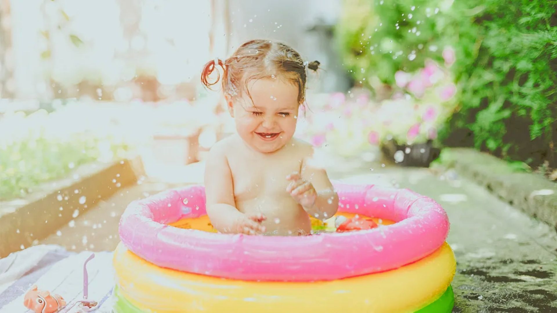 A smiling toddler splashes in a small inflatable pool with pink and yellow rings on a sunny patio.