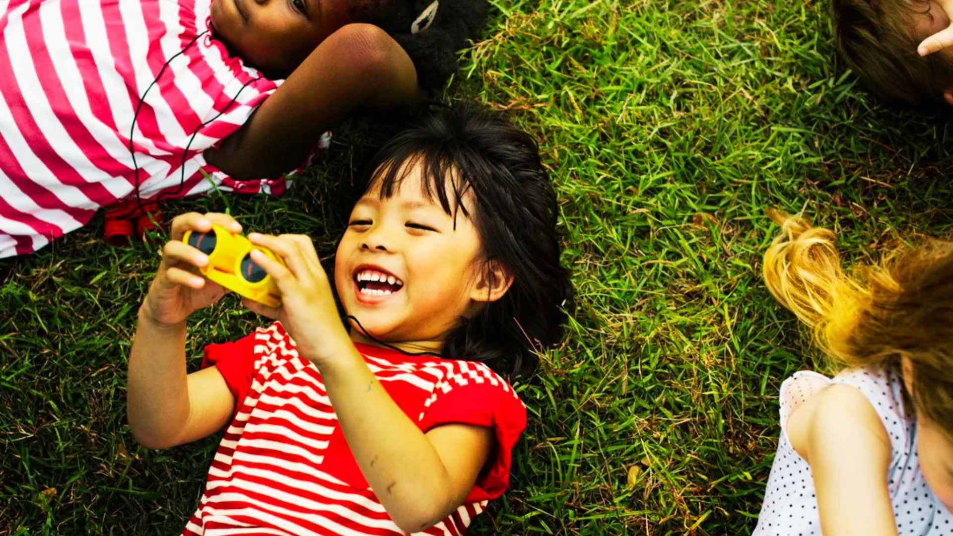 Children lying on grass, engaged with cell phones, highlighting the balance between technology and outdoor play.