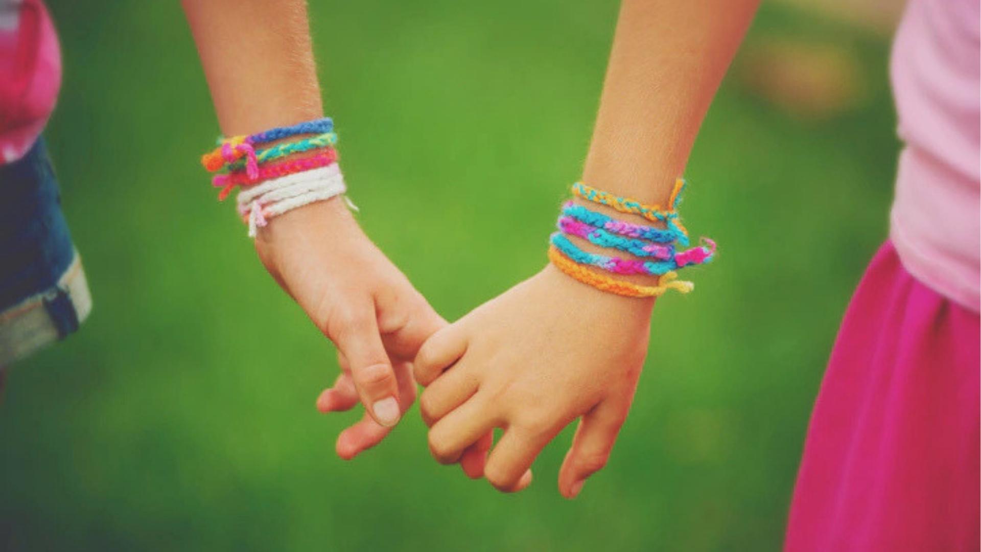 Two children holding hands, colorful woven friendship bracelets on wrists, green grass background.
