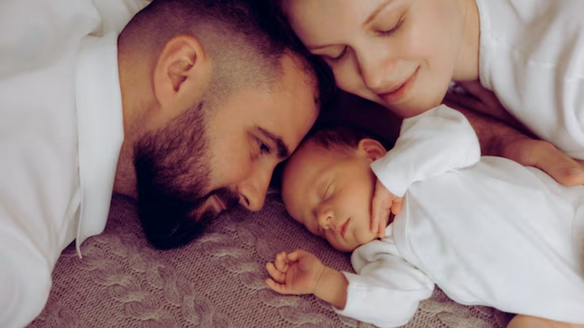 A man and woman lie on a bed, gently cradling a sleeping baby between them.