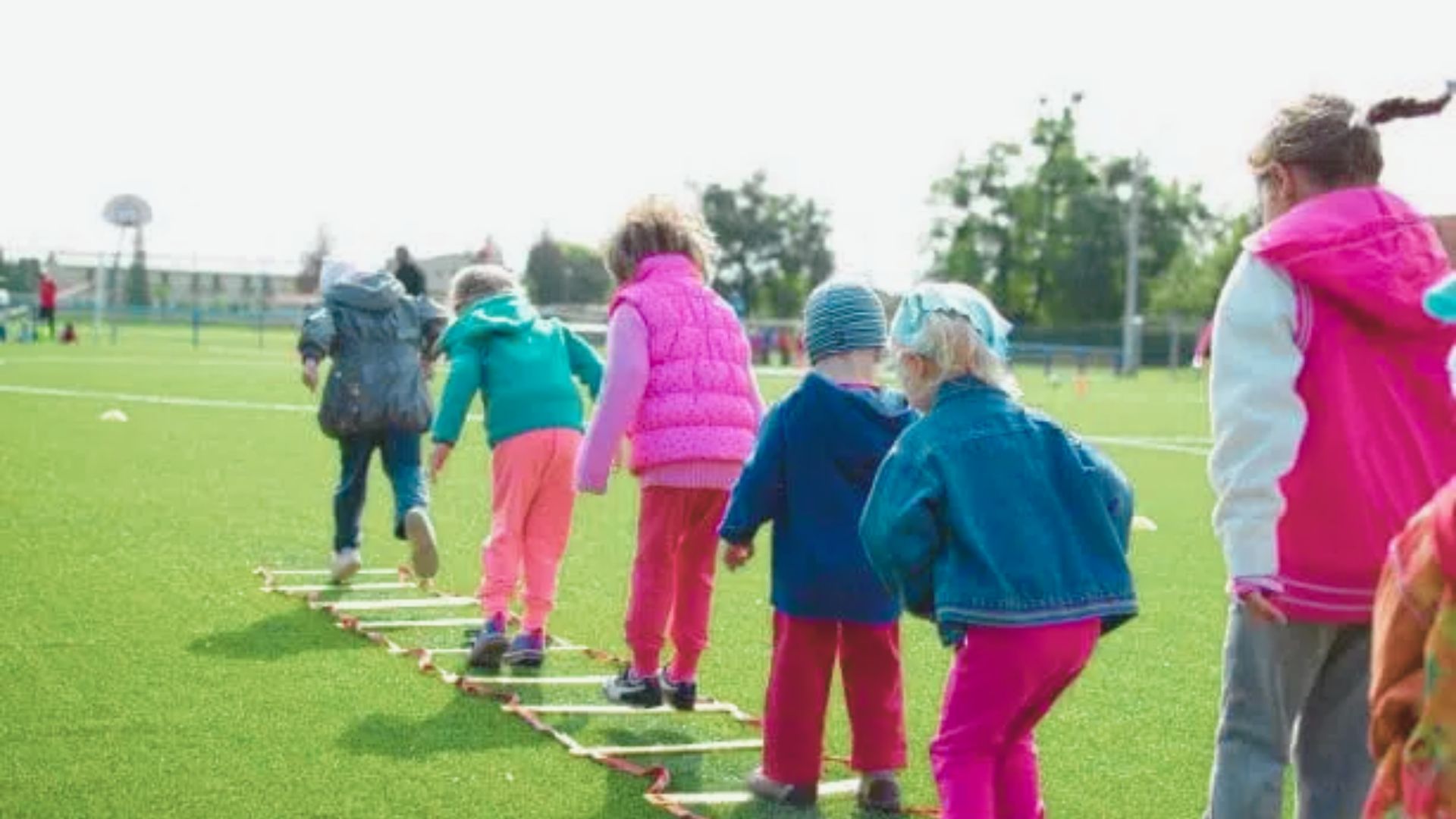 A line of children in bright jackets step through an agility ladder on a green field during an outdoor game.