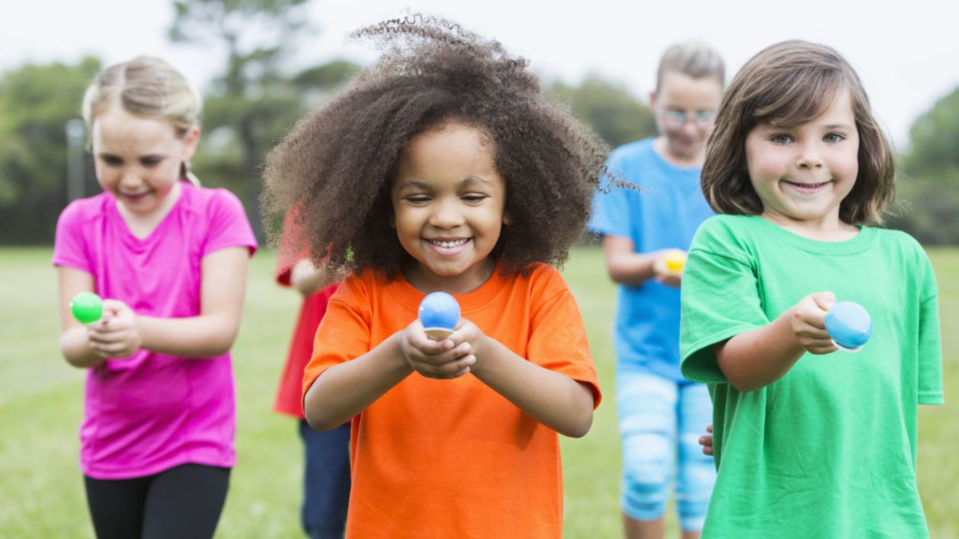Smiling kids outdoors playing an egg and spoon race on grass, a fun farm field day activity for home or school