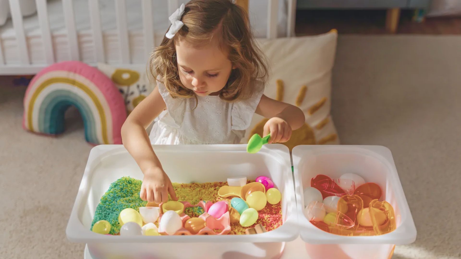 A toddler in a white dress finds a sensory bin filled with colorful rice and plastic eggs using a small spoon.
