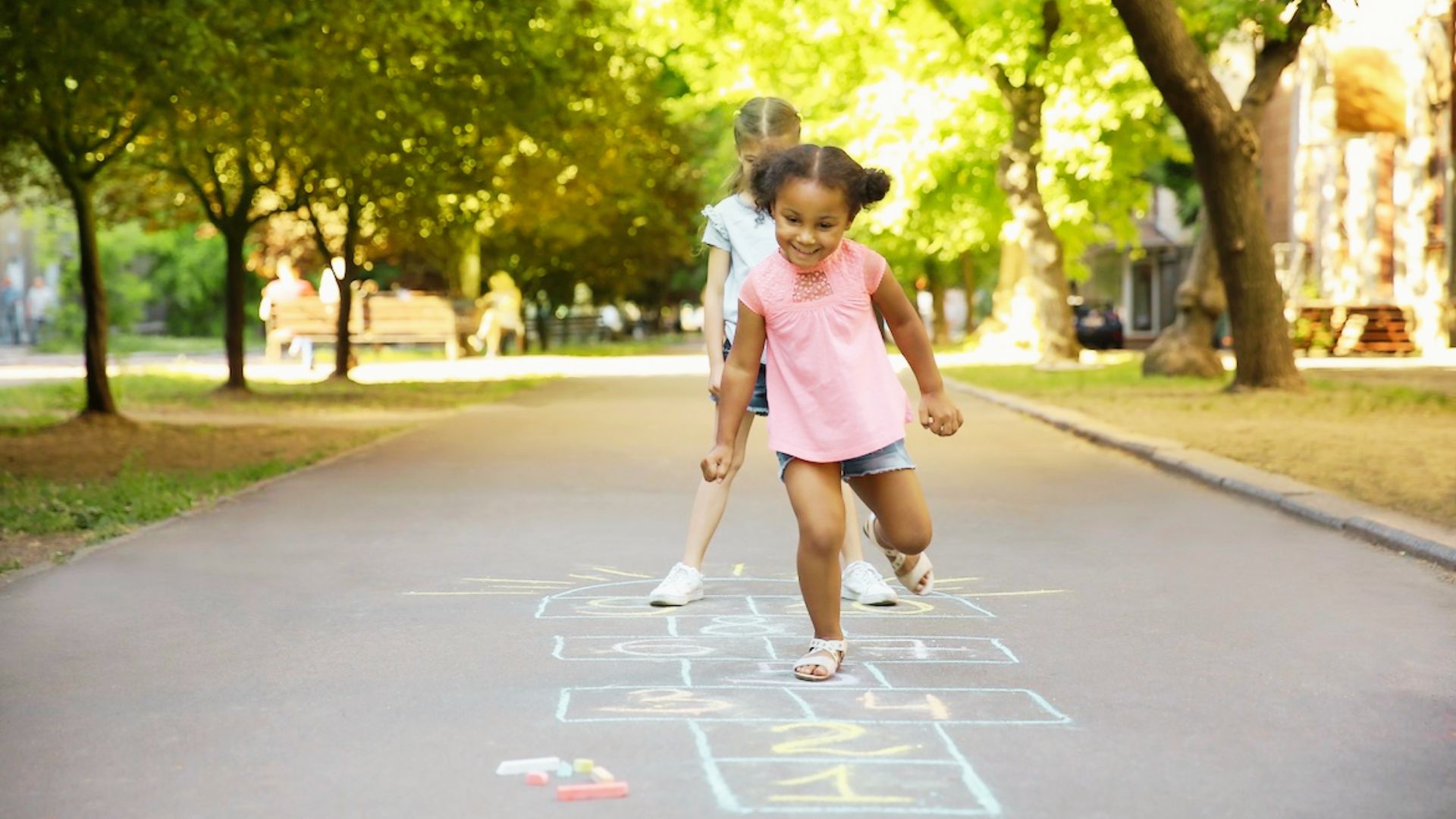 Two children joyfully playing hopscotch on a sunny sidewalk, surrounded by colorful chalk drawings.