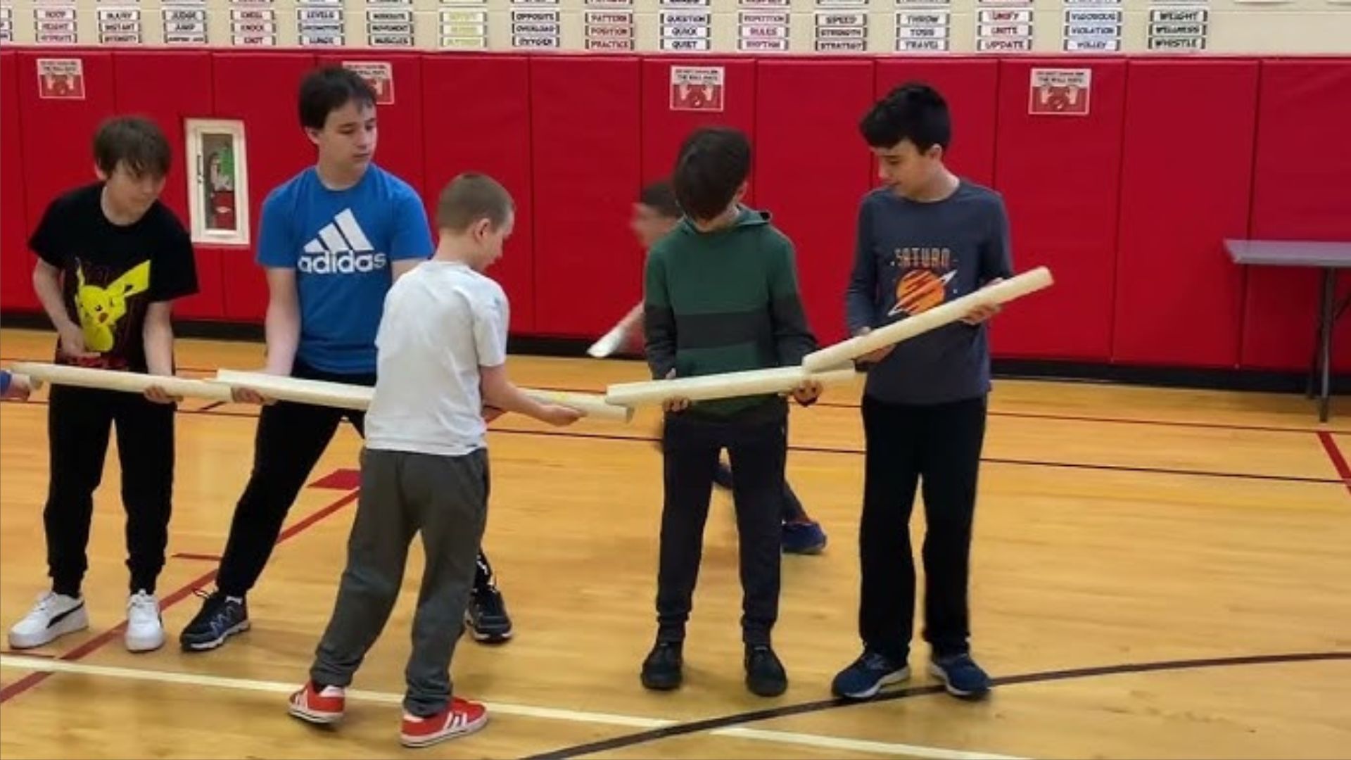 Group of five boys in a school gymnasium holding long pipe together, participating in a cooperative team-building physical activity for kids at school