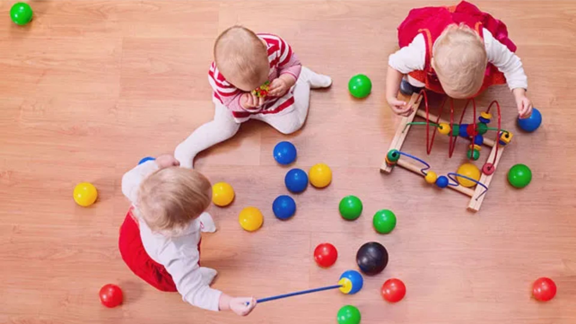 Three toddlers sitting on the floor sharing colorful balls and a bead maze toy, developing parallel play and early sharing skills as a friendship activity for preschoolers