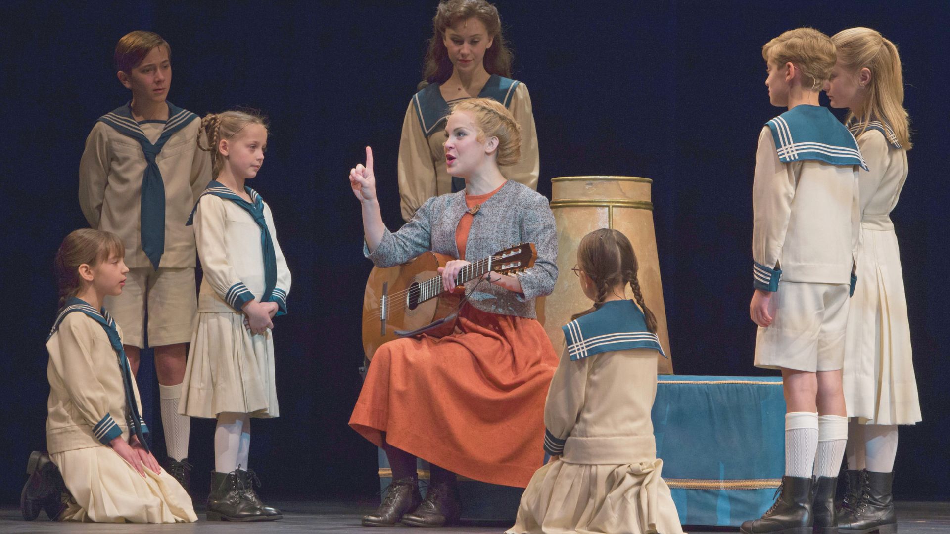 A woman plays an acoustic guitar while teaching a group of children in sailor costumes during a stage performance.