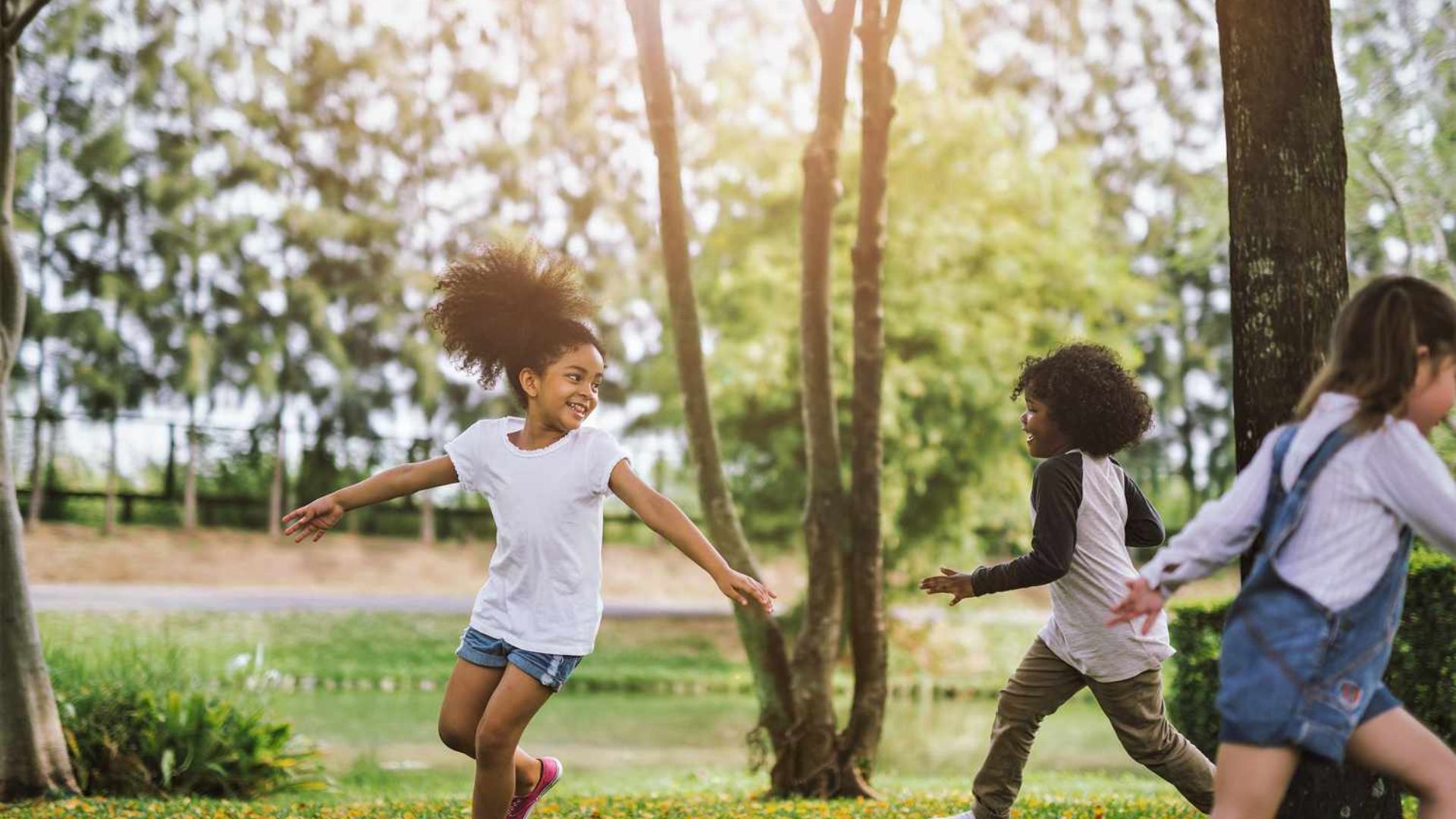 Three preschool friends running and playing a chasing game in a park, laughing together while building gross motor skills and friendship bonds for preschoolers