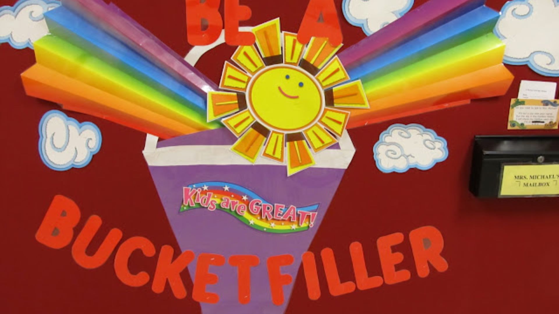 Vibrant classroom bulletin board with a purple bucket, rainbow rays, smiling sun, and large text reading “BE A BUCKET FILLER.”