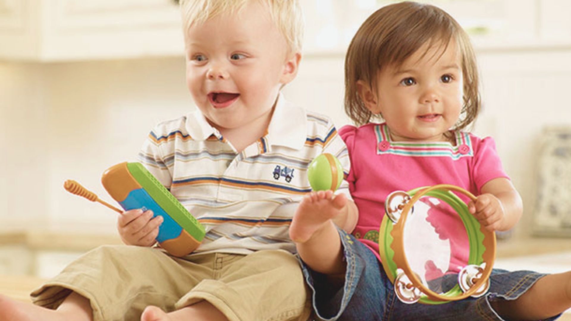 Two toddlers sit together holding musical toys, one with a tambourine and the other with a maraca.