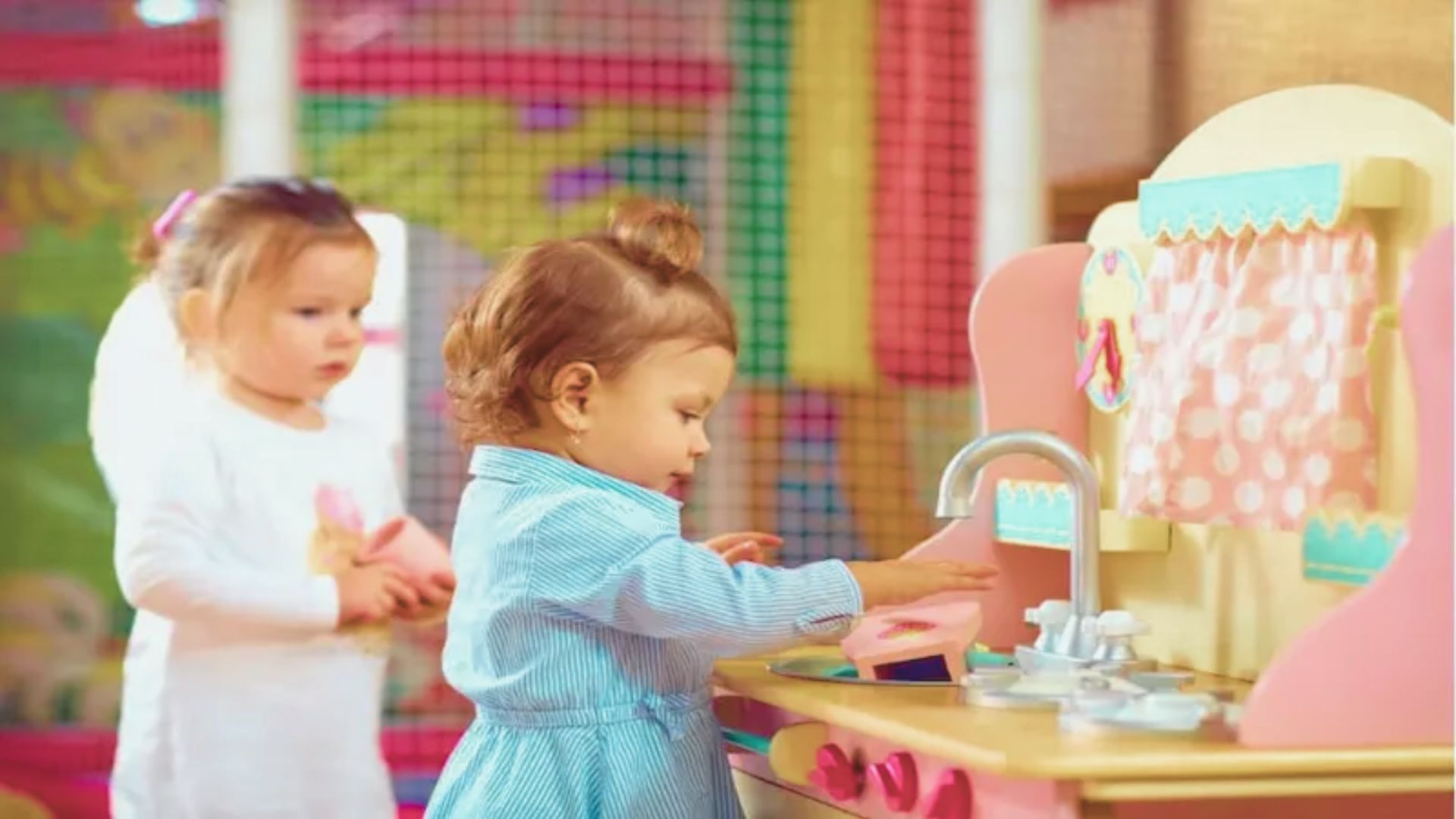 Two toddlers engage in pretend play at a colorful toy kitchen.