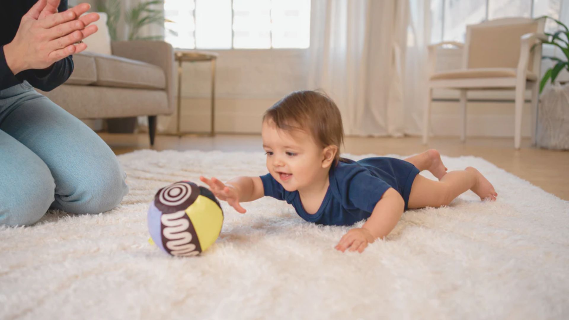 An infant in a navy onesie lies on a white rug, reaching for a high-contrast patterned ball as an adult claps.