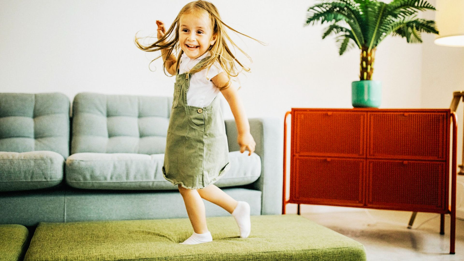 A little girl is playing “floor is lava” joyfully jumps on a couch in a cozy living room filled with soft lighting and colorful decor.