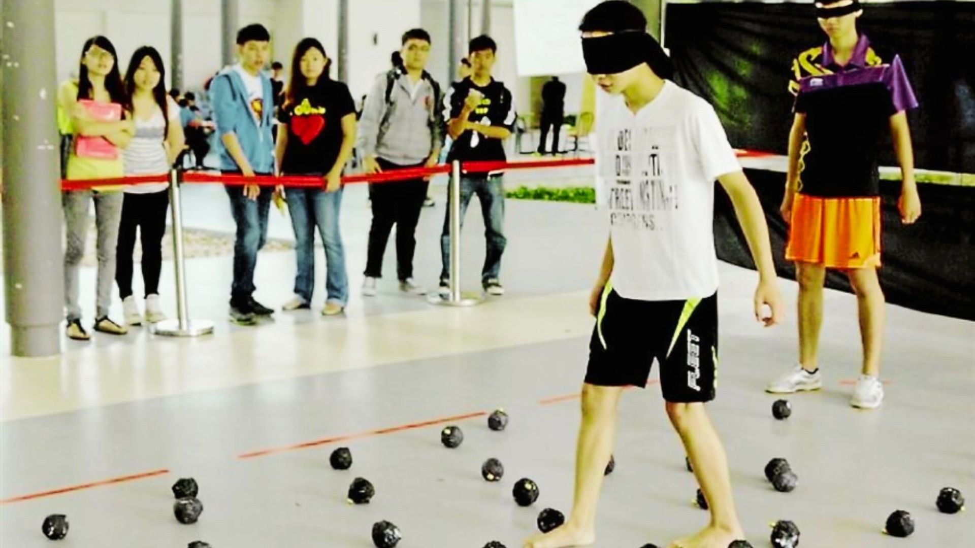 A man stands in front of a group of blind folded attentive people, engaging them with his presentation. With balls placed everywhere on the floor.