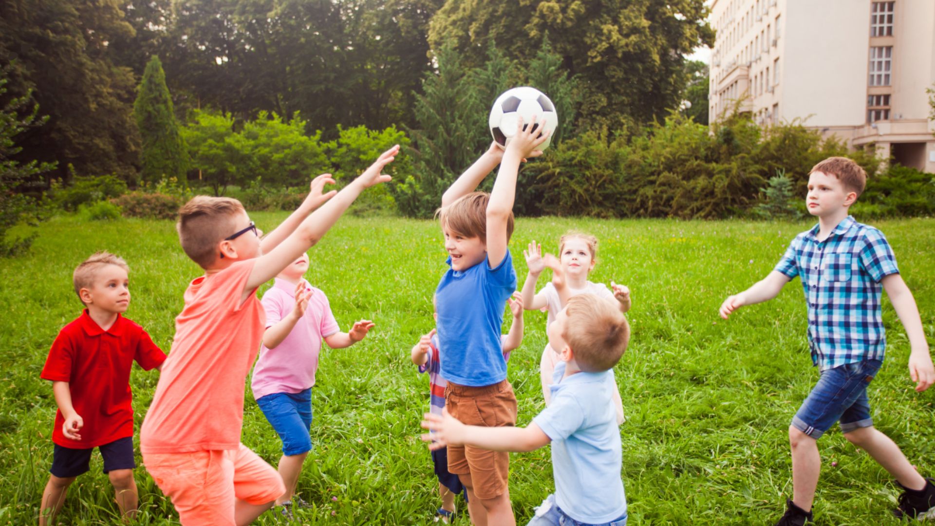 Group of seven kids playing with a soccer ball on a grassy field, reaching and taking turns.
