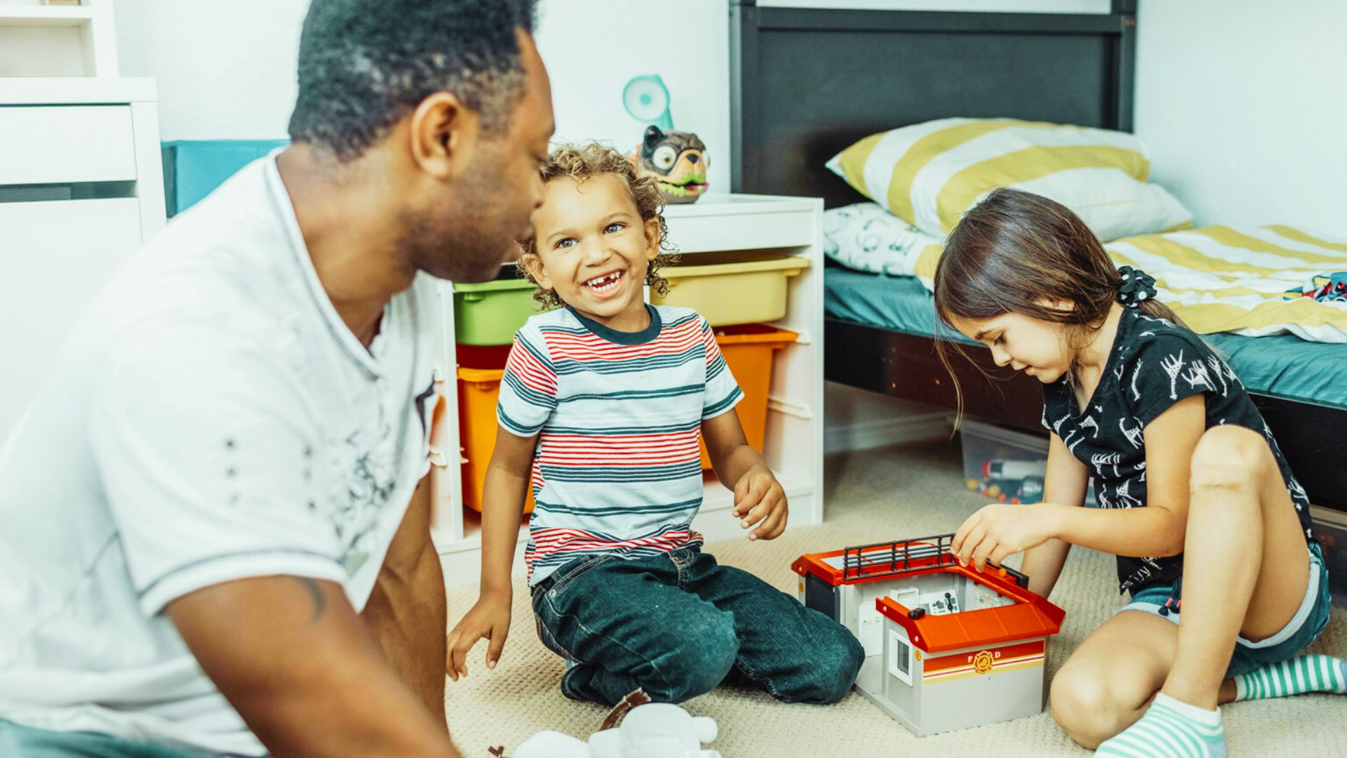 A father and his children joyfully playing with a colorful toy box in a cozy bedroom.