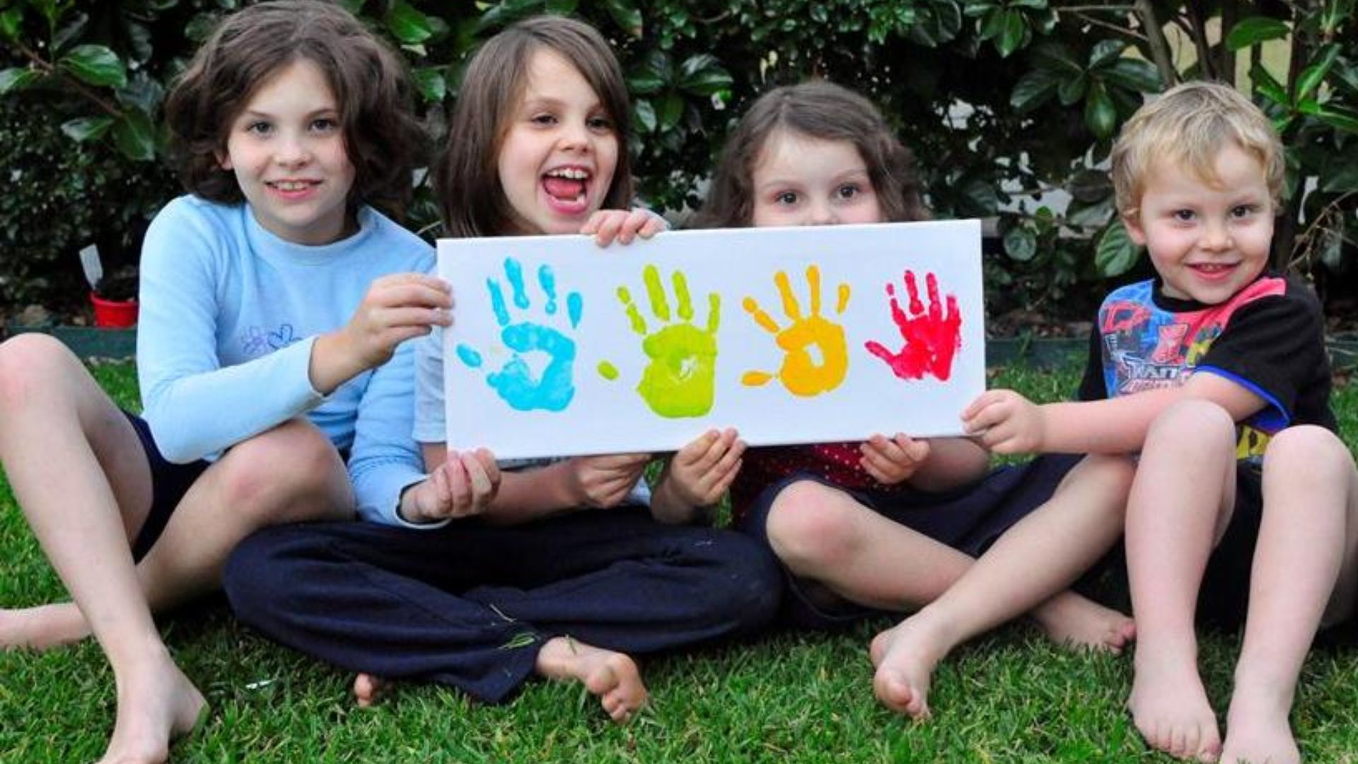 Four smiling preschool children sitting on grass holding a canvas with colorful handprints, celebrating teamwork and shared art as a friendship activity for kids