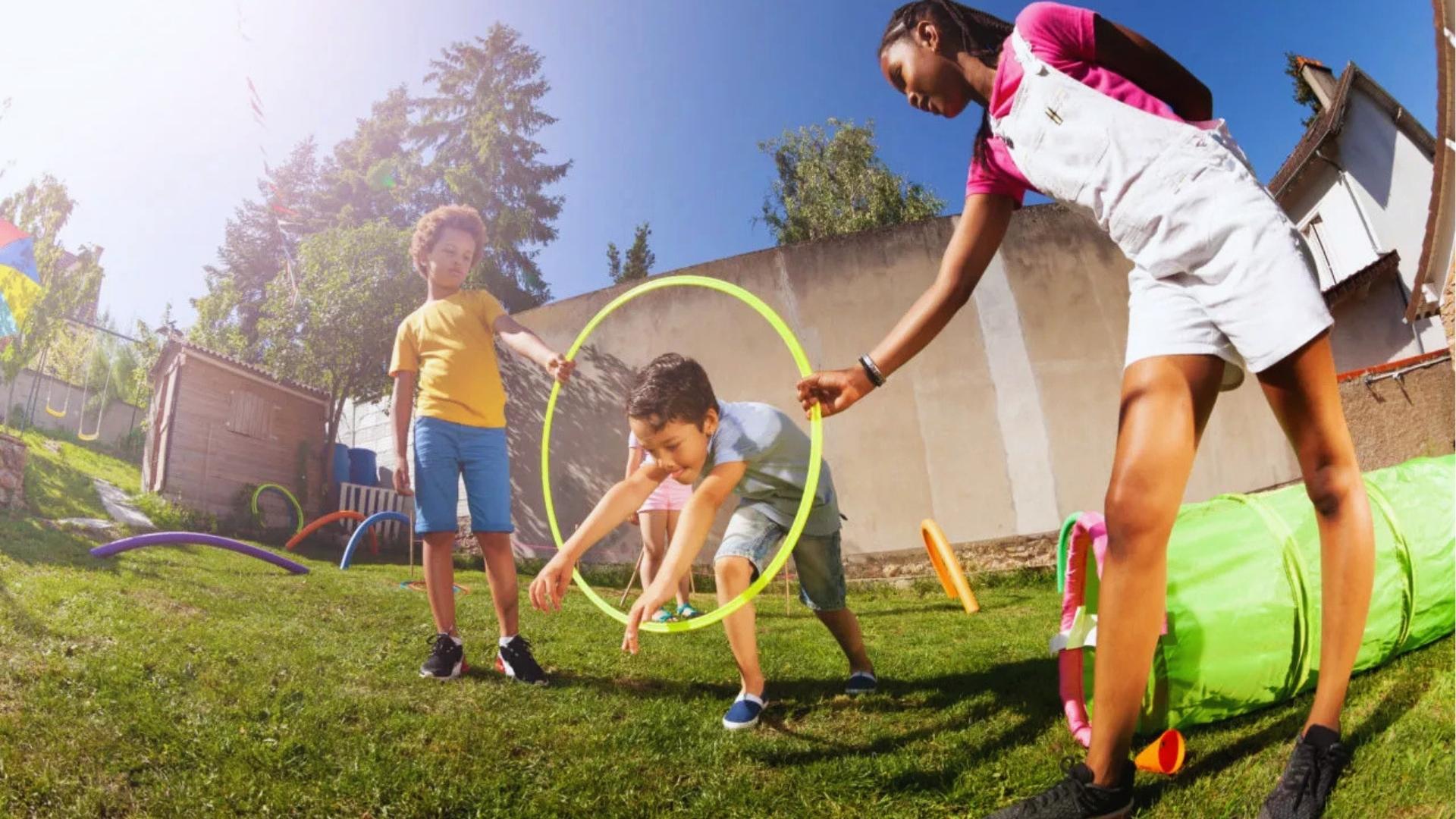 Kids play outdoor obstacle course, boy crawls through hula hoop, sunny backyard.