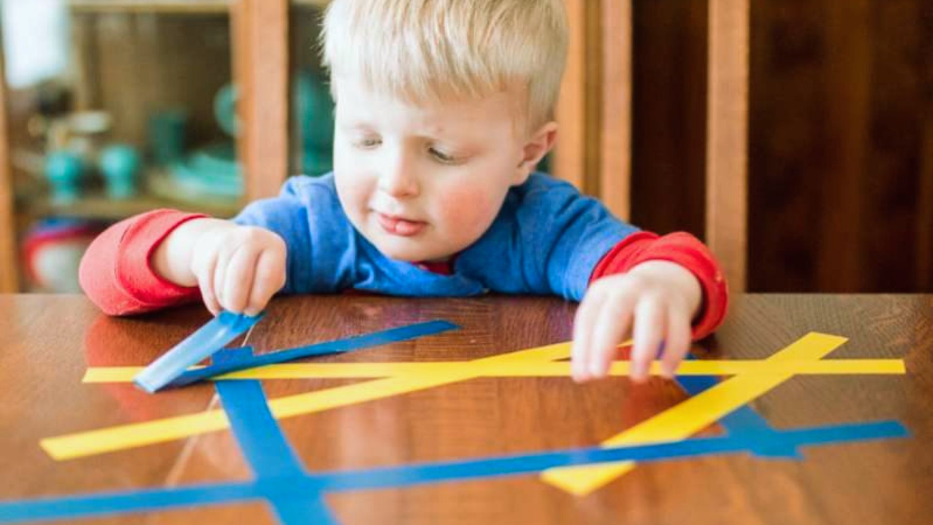 A focused toddler in a blue shirt peels blue and yellow tape strips off a wooden table.