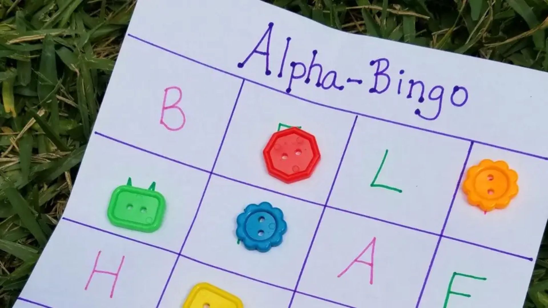 Hand-drawn Alpha-Bingo card on white paper with purple grid lines, letters B, L, H, A, F written in markers, and colorful plastic buttons covering some squares, placed on grass.