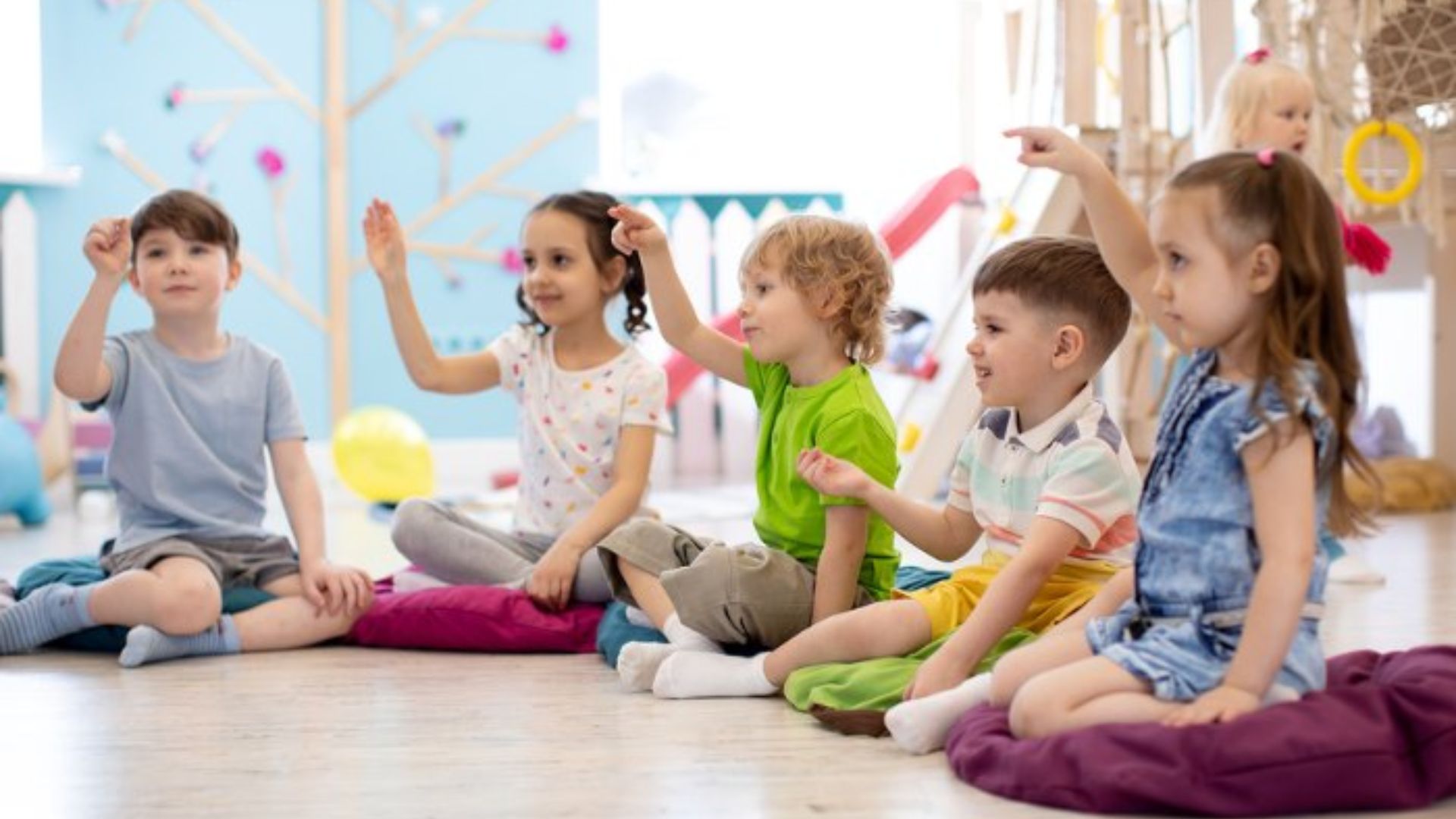 Five preschool kids sitting on floor cushions in a classroom, raising hands and participating in a group discussion circle time activity at school