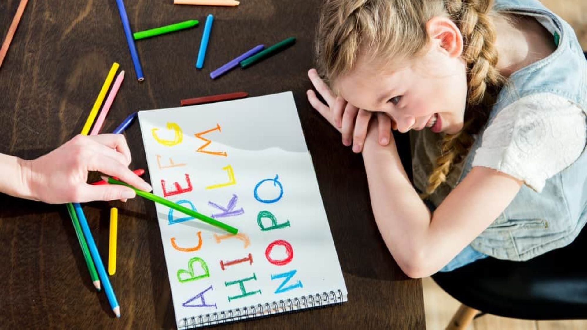 A young girl with braids smiles at a spiral notebook filled with colorful handwritten alphabet letters.