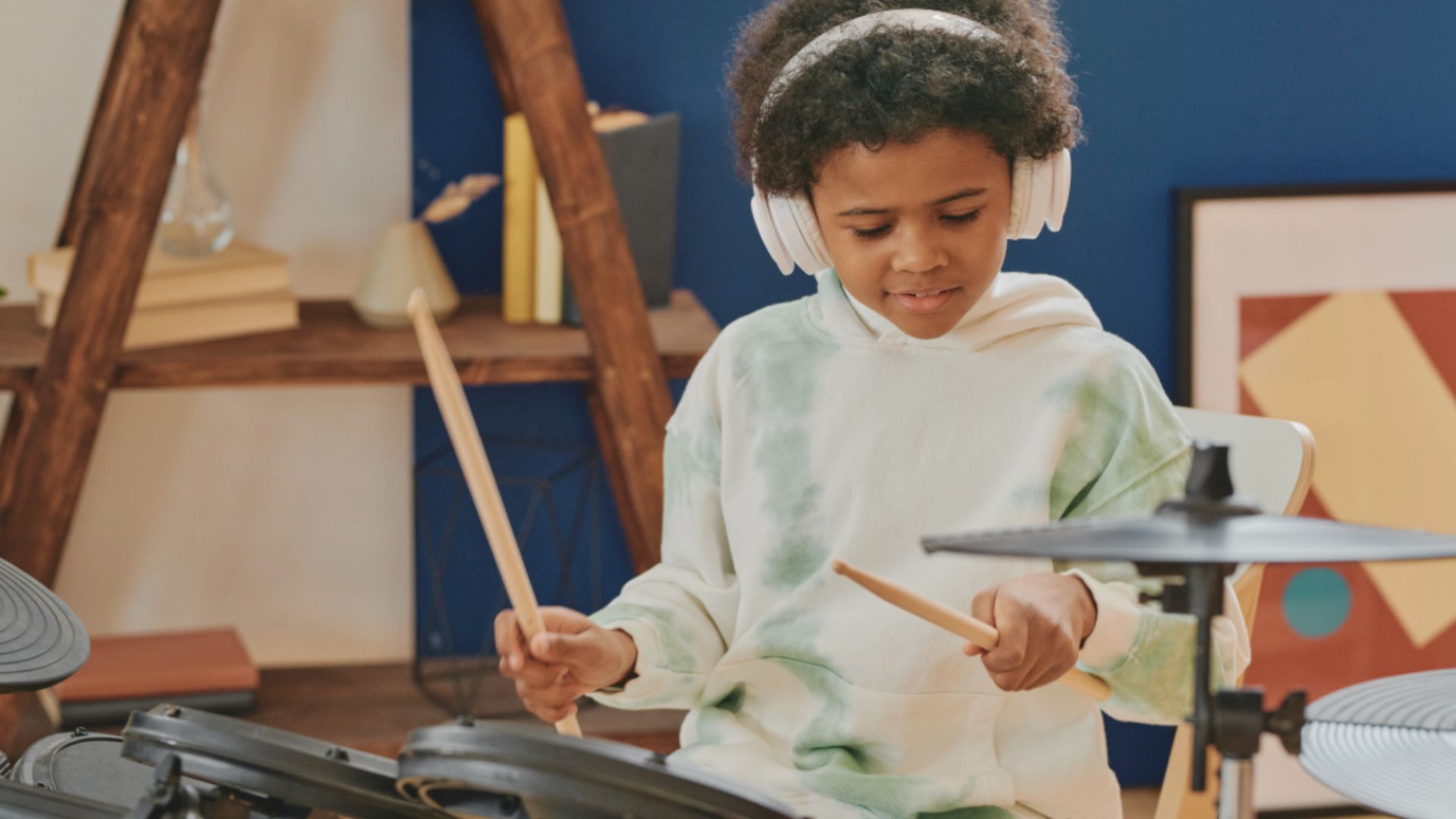 A child wearing white headphones concentrates while playing an electronic drum kit with drumsticks.