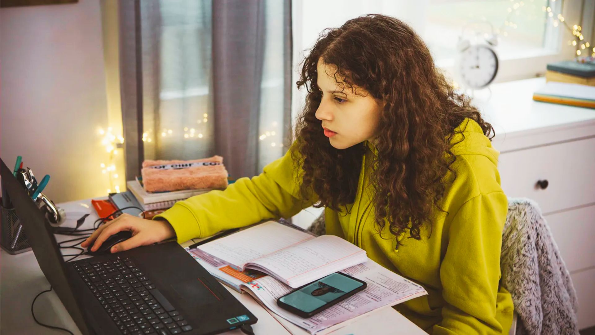 A girl sitting at a desk, focused on her laptop, surrounded by books and stationery, engaged in problem-solving activities.