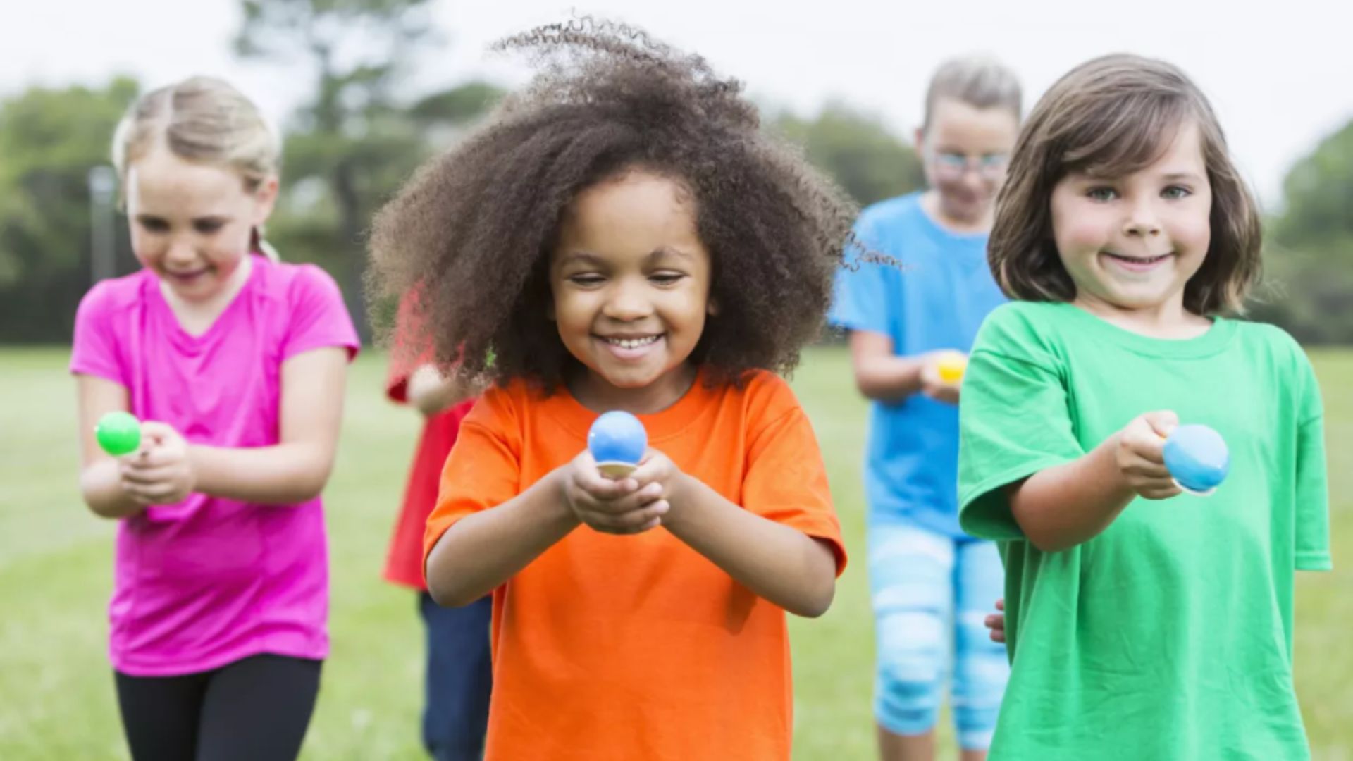 Smiling preschool children playing an outdoor egg and spoon race game together, developing sportsmanship and peer encouragement through a friendship activity for kids