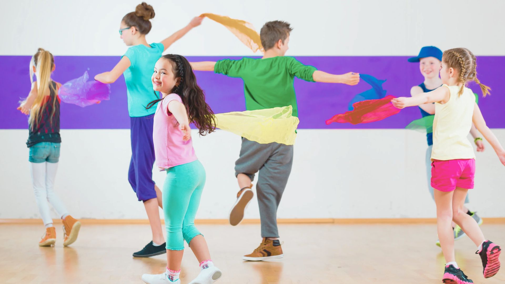 Children wave colorful scarves while dancing in a bright studio with a purple wall.
