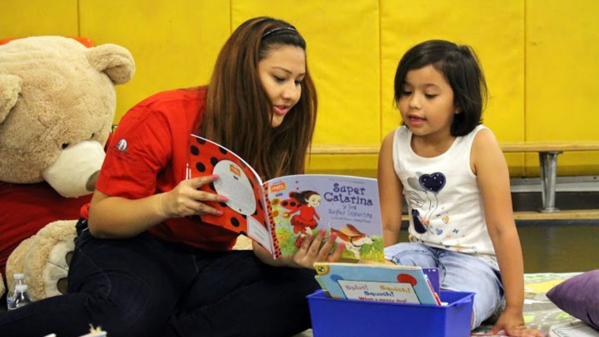 Teacher reading a colorful storybook to a preschool girl on the floor, modeling peer connection and shared attention as a friendship activity for preschoolers