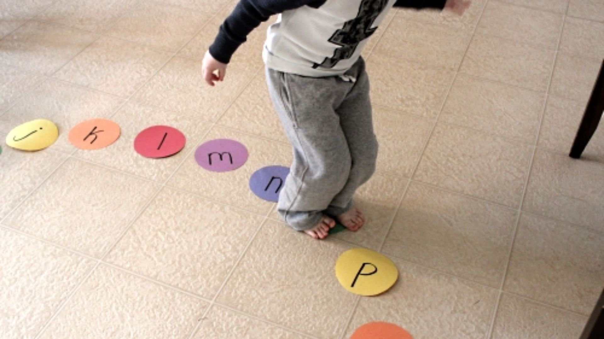 Barefoot child in gray sweatpants hopping on colorful paper circles with lowercase letters j, k, l, m, n, p arranged on tiled floor.