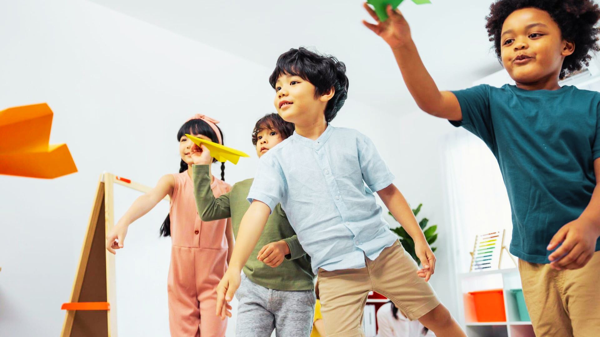 Children joyfully playing with colorful paper airplanes in a bright classroom filled with desks and educational posters.